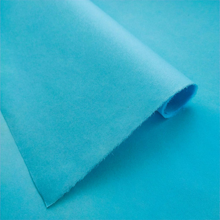 Sheets of solid-colored Kozo Studio mulberry paper in Atoll Blue with delicate texture and curled corners on a light blue surface.