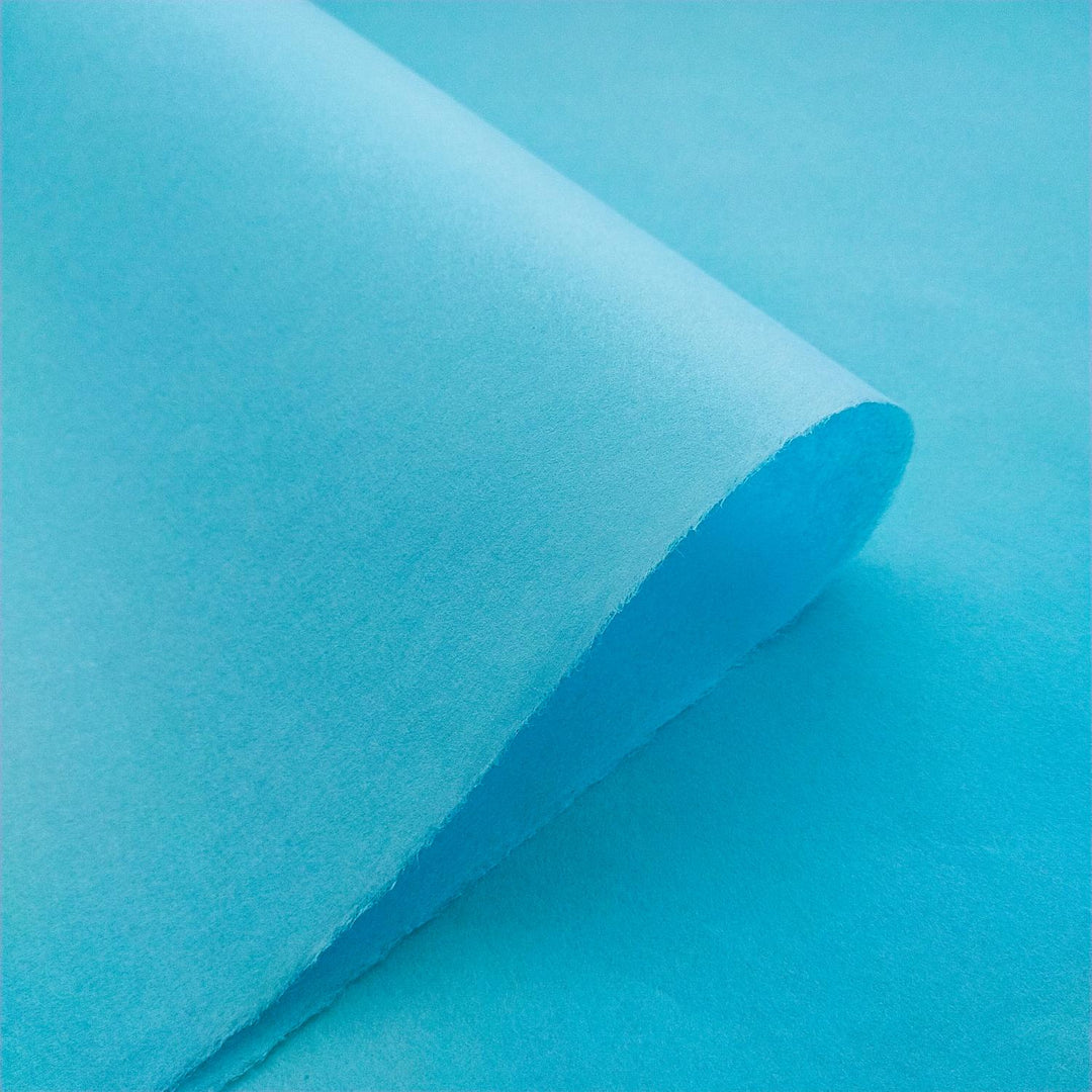 A close-up of partially curled Solid-Colored Kozo Paper (Atoll Blue) by Kozo Studio against a matching blue background. The smooth, evenly colored texture subtly contrasts with the slightly different shade of the background.