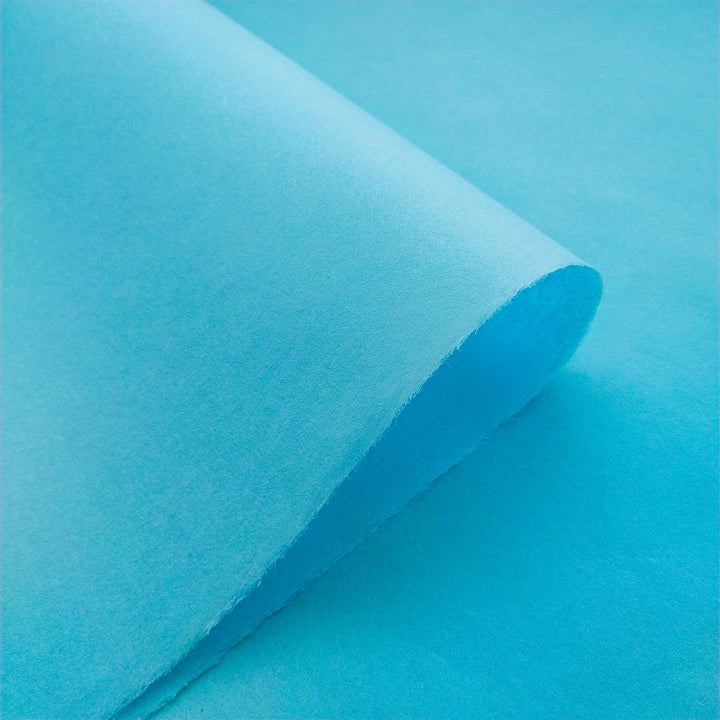 A close-up of partially curled Solid-Colored Kozo Paper (Atoll Blue) by Kozo Studio against a matching blue background. The smooth, evenly colored texture subtly contrasts with the slightly different shade of the background.