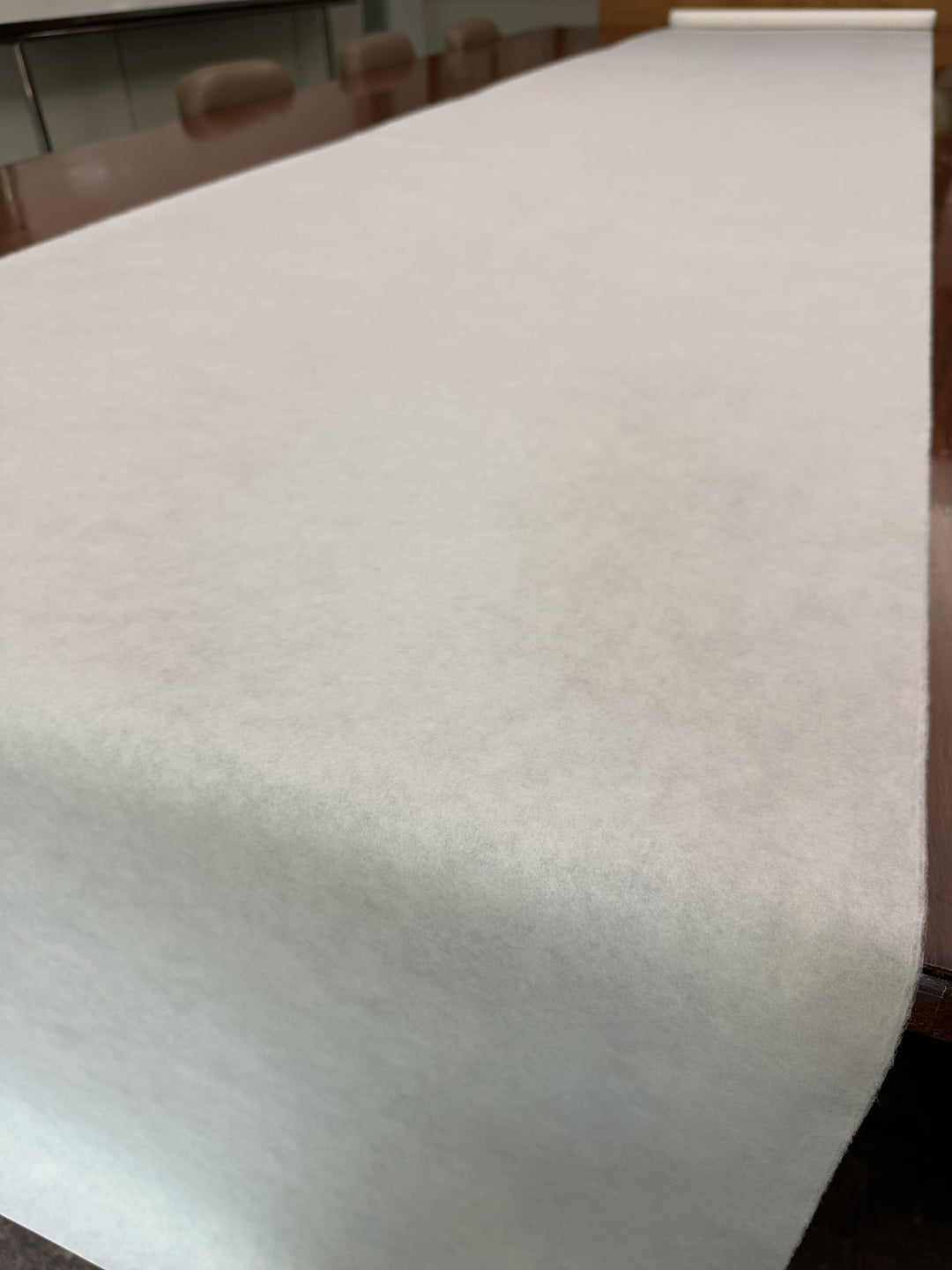 Eine dünne weiße Kozo-Papierrolle (45 g/m²) von Kozo Studio wird entlang eines großen, polierten Konferenztisches, umgeben von Stühlen, in einem Besprechungsraum abgerollt.